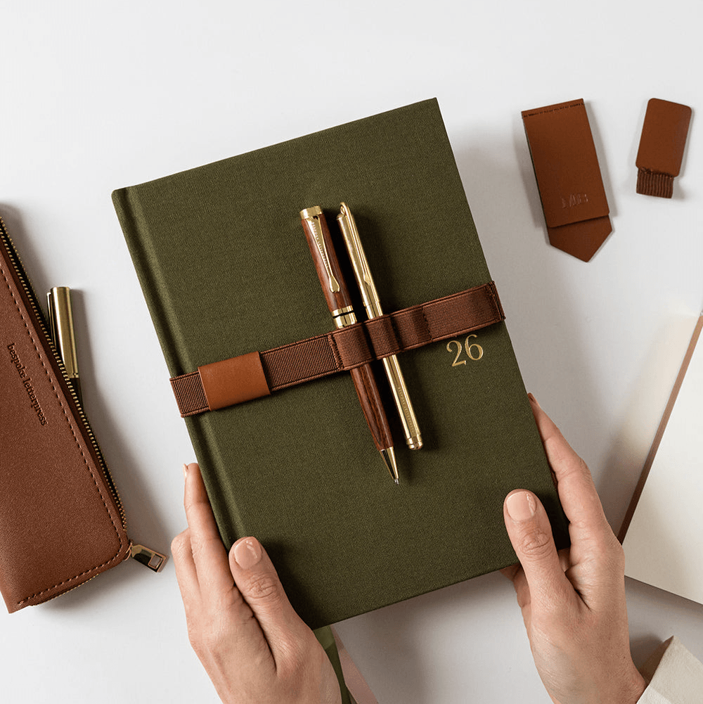 Hunter Green notebook with a brown elastic book strap and 2 pens, held by hands on a light background along with brown bookmark, pen loop, tan slim pencil case.