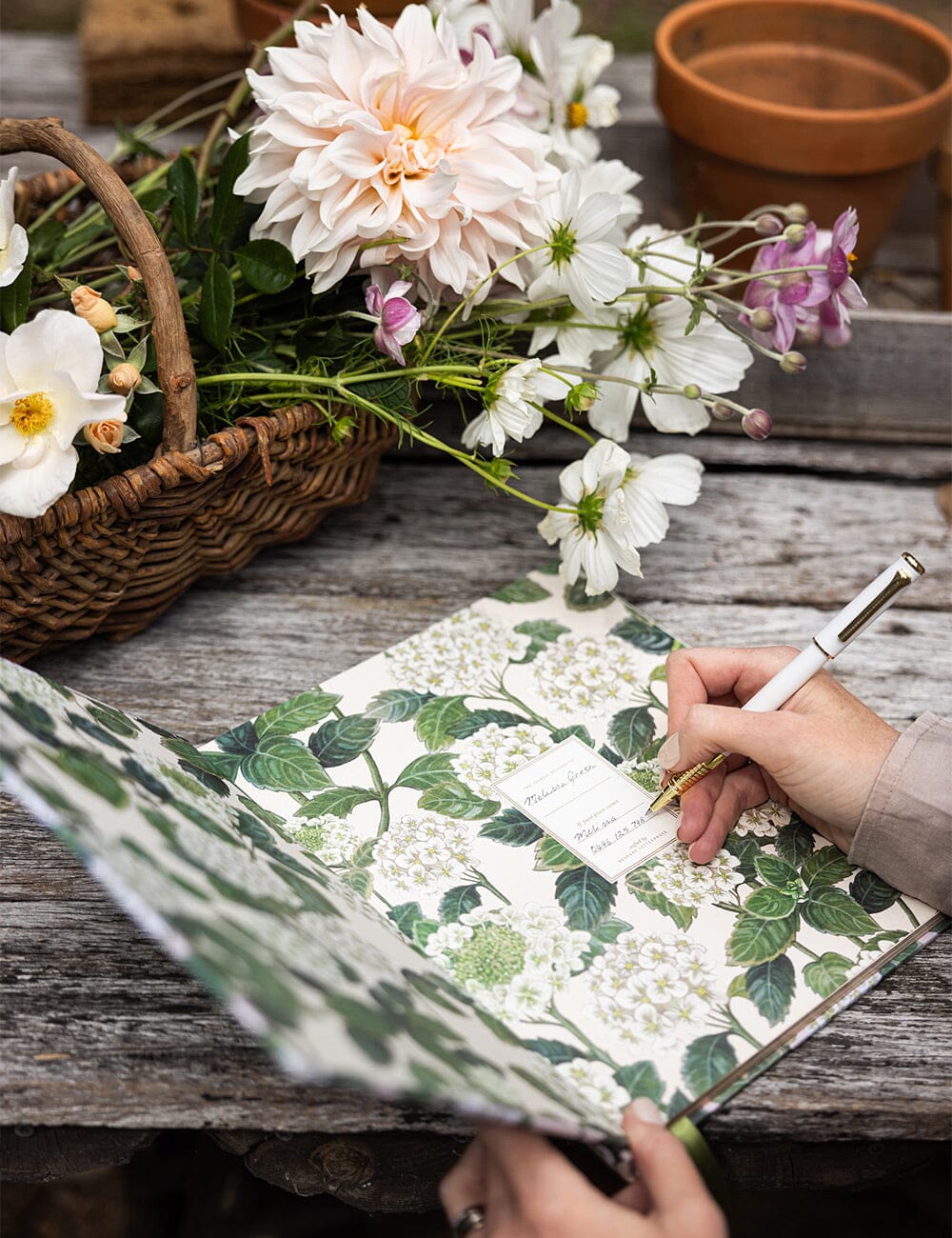 Hand writing a name inside the Gardening Journal – Hydrangea, set on a rustic wooden table beside a basket of freshly picked blooms and garden tools.