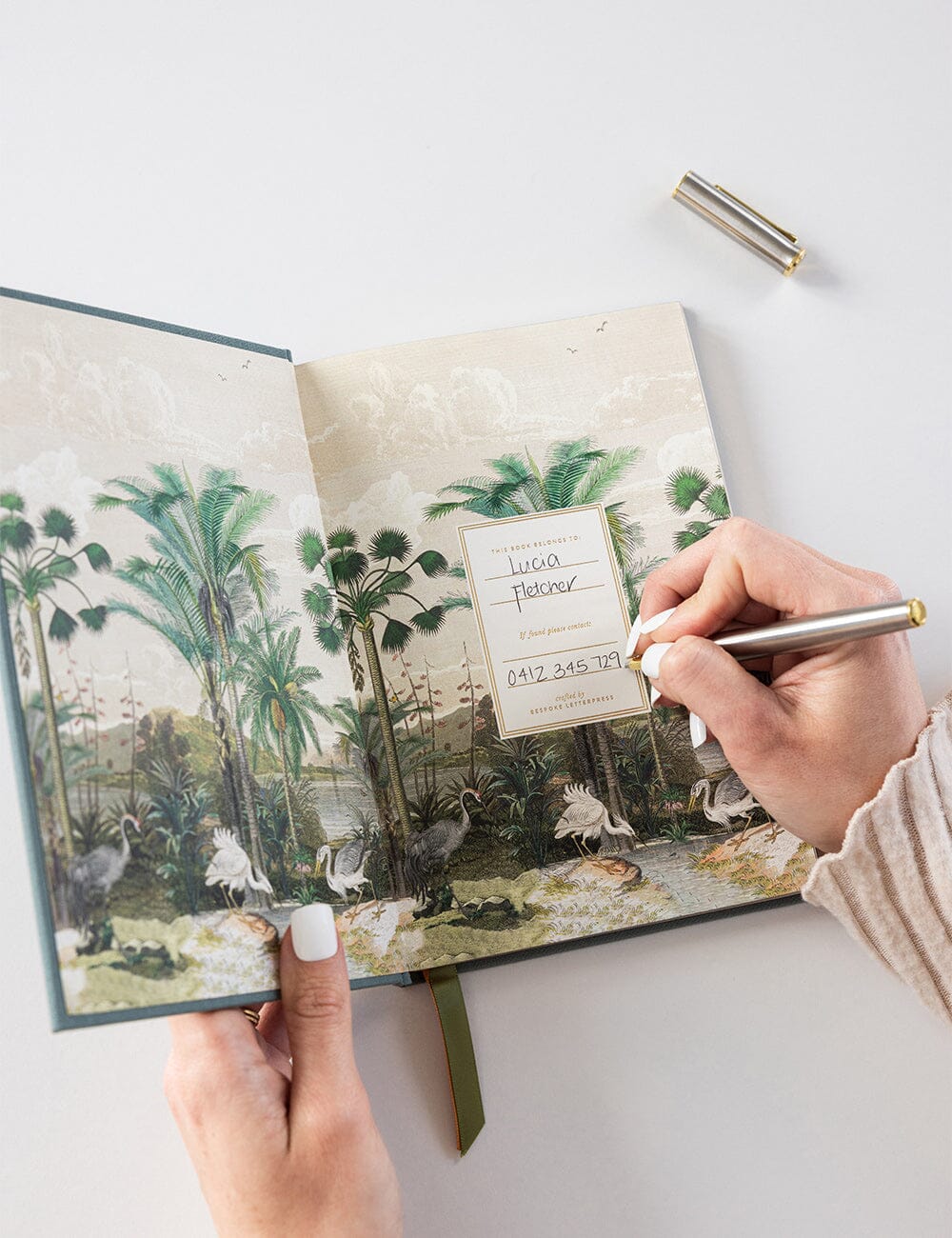 A hand writing in the name and contact details on the ownership page of the Bespoke Letterpress Gratitude Journal in Seaglass. The inside cover features a lush, vintage-style illustrated landscape with exotic birds and palm trees in soft, muted tones.