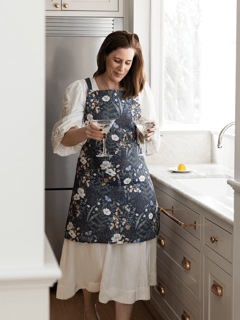 Woman wearing a floral linen apron by Bespoke Letterpress in a kitchen, featuring a dark botanical print and premium designer kitchen linen.