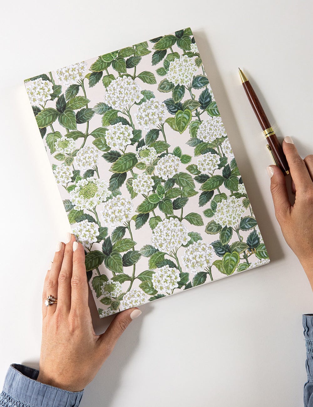 Hands holding the Bespoke Letterpress Hydrangea Jumbo Notepad Jotter on a white desk, showcasing its elegant botanical cover featuring white hydrangea blooms and lush green leaves.