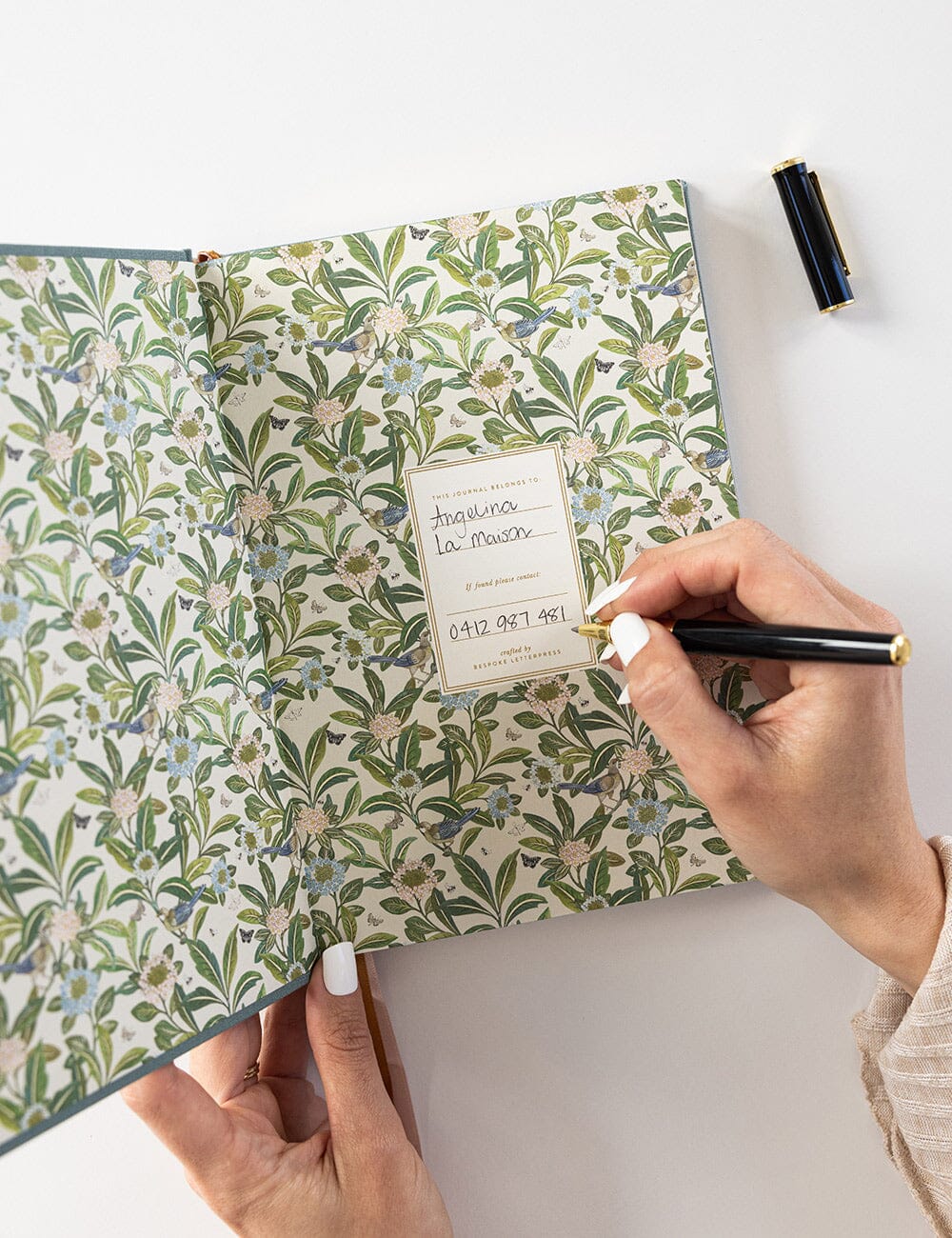 Person writing inside the Seaglass Linen Bound Journal, showing the floral patterned endpapers and nameplate page with 'This journal belongs to' label.