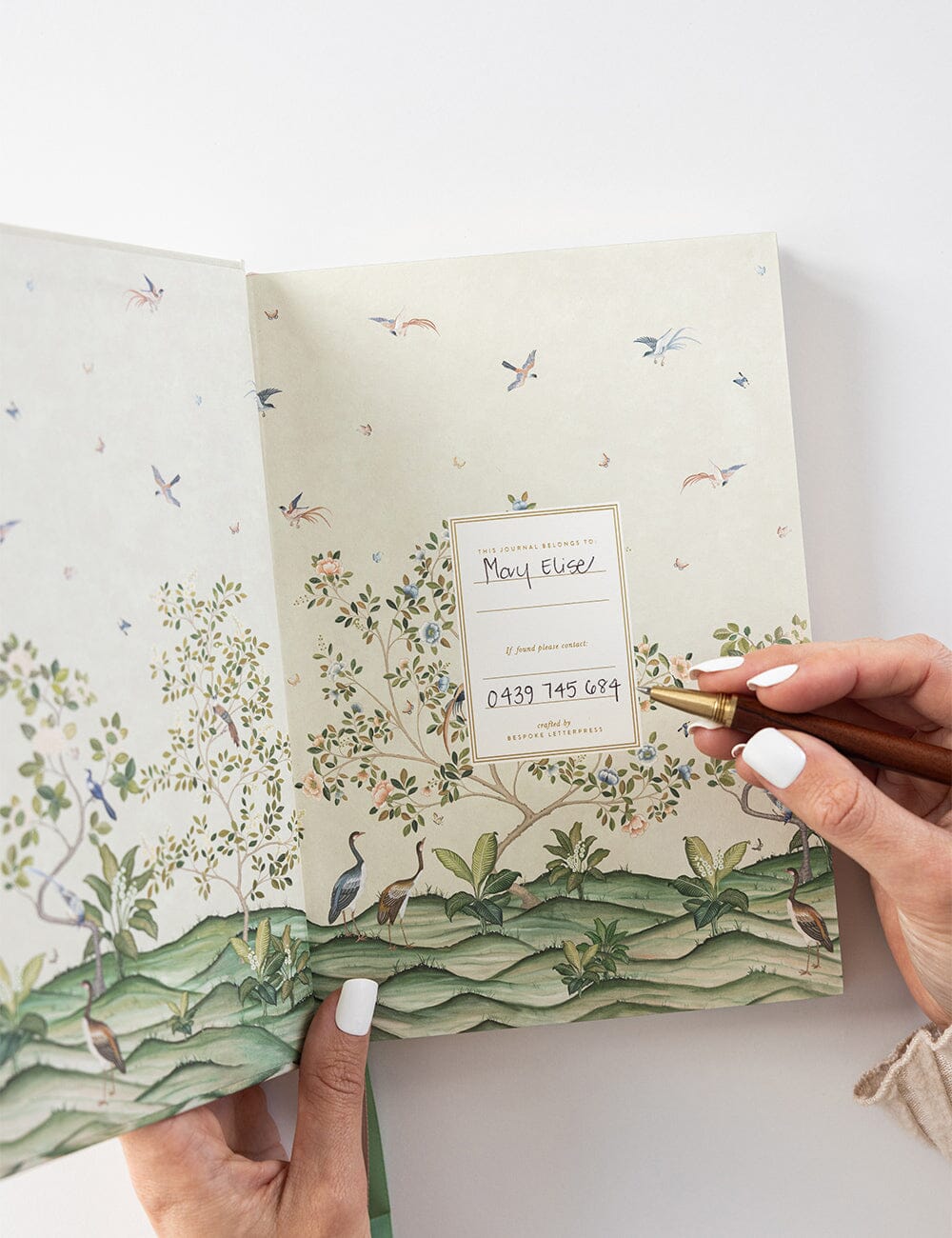 Person writing contact details on the “This Journal Belongs To” page of the Spring Landscape journal, featuring an illustrated nature scene with birds and trees.