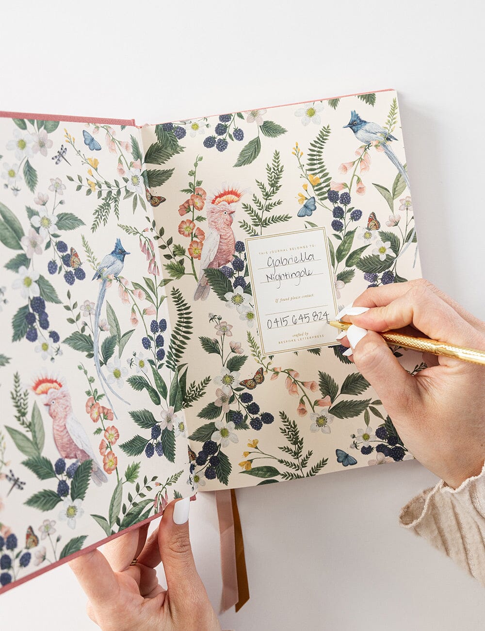 Person writing their name in the Tea Rose Linen Bound Bullet Journal, featuring illustrated botanical endpapers with native birds, blackberries, and florals.