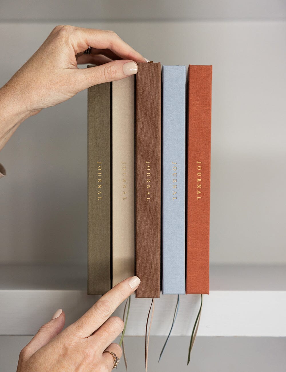 Five journals in earthy tones of brown, green, beige, blue and terracotta are stacked on a bookshelf with a hand holding the middle journal. Each journal features a gold foiled title on the spine with the satin ribbon markers peeking out over the shelf for an added touch of luxury.