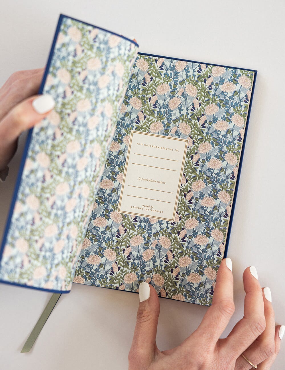 Close-up of the inside front cover of the Royal Blue Soft Cover Notebook, revealing a beautifully patterned floral endpaper in soft blue, green, and blush hues. A gold-edged nameplate for personalisation is centered on the page, with a hand gently holding the cover open.