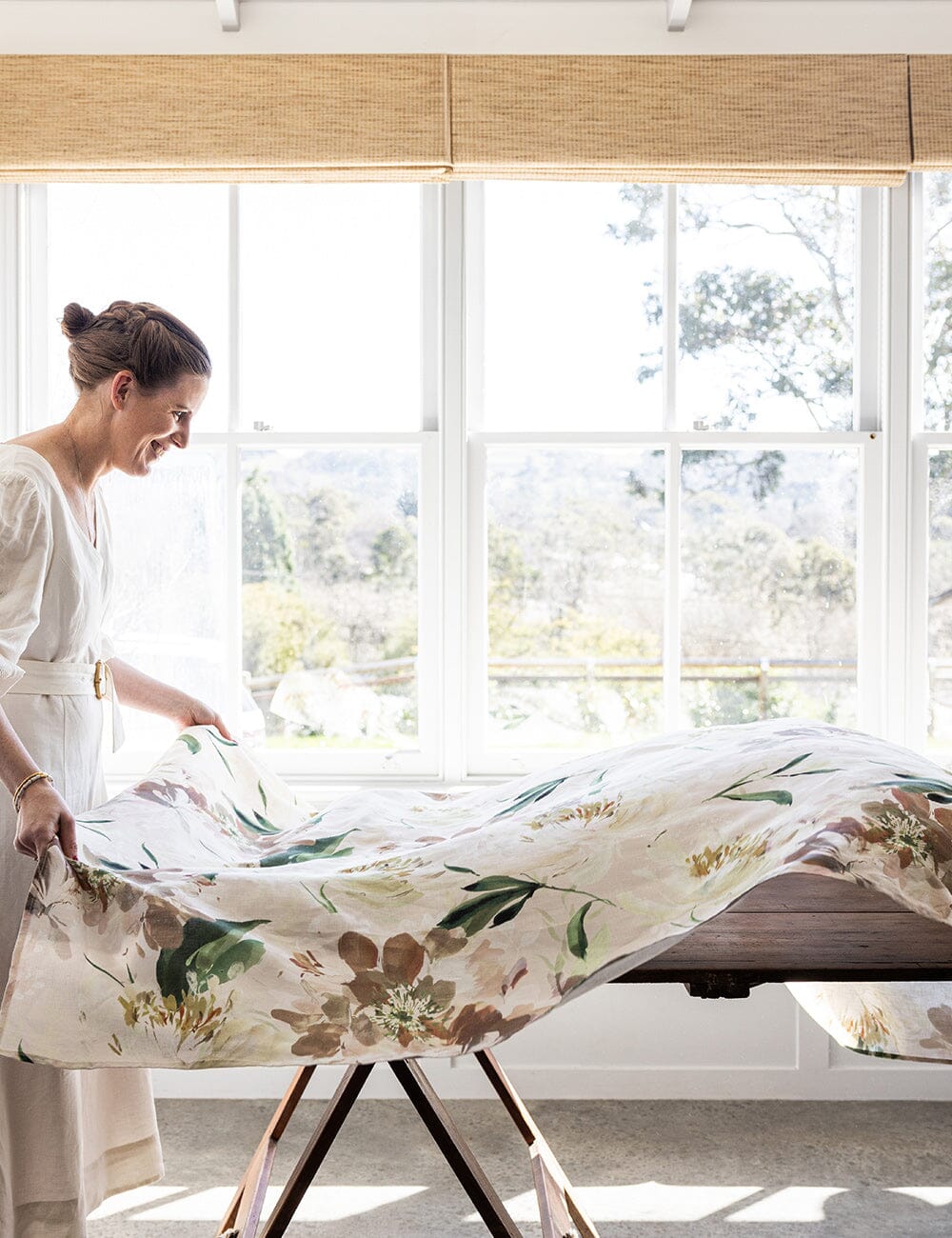 Woman styling a dining table with the designer Peony Study linen tablecloth, featuring a floral print in a sunlit room with large windows.