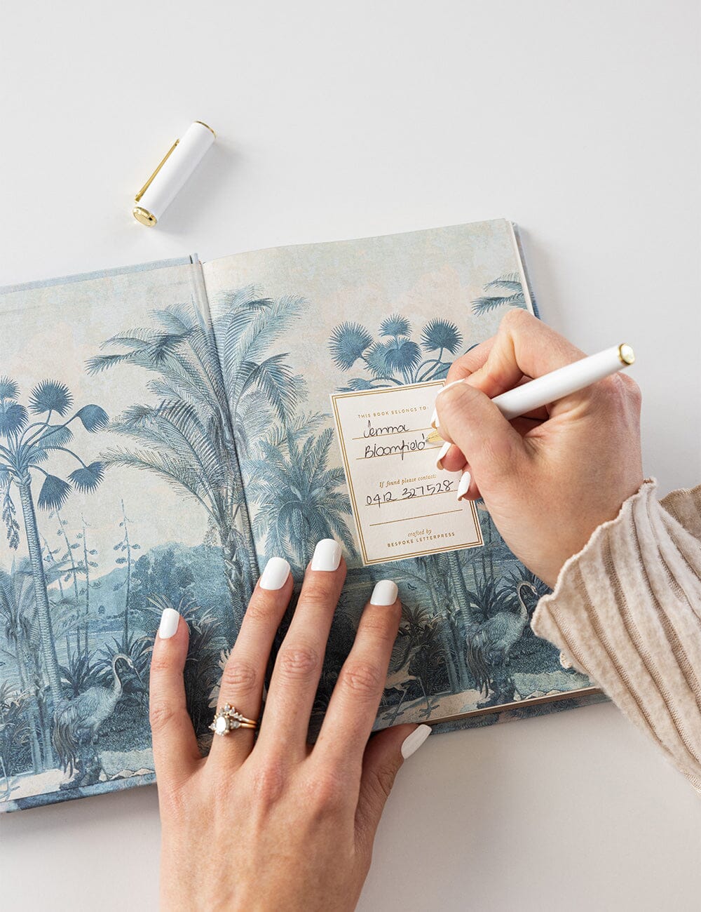 Close-up of a hand writing a name and contact number on the “This Book Belongs To” page inside the Bespoke Letterpress Travel Planner Journal, featuring a scenic blue tropical illustration on the endpaper.