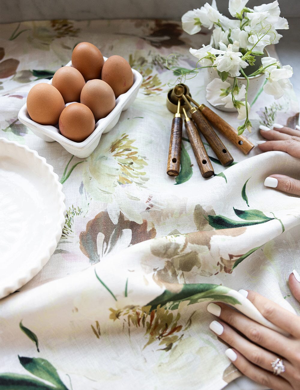 Peony linen tea towel styled on a kitchen bench with fresh eggs, measuring spoons, white sweet pea flowers, and soft floral artwork on 100% flax linen.