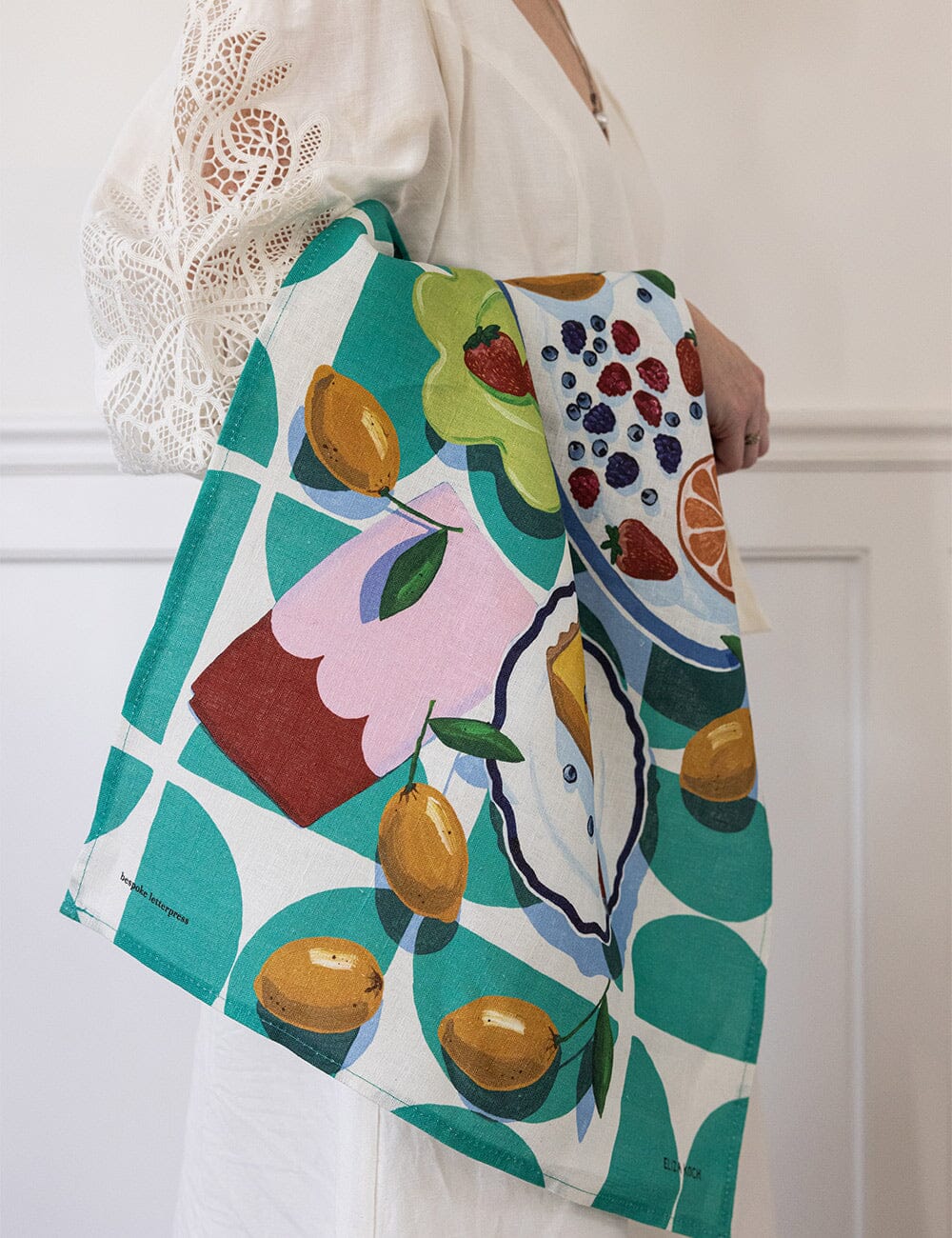 A woman in a white embroidered blouse holds the Lemon Tart & Berries Linen Tea Towel draped over her arm, showcasing its lively illustration of fruit and desserts in vibrant tones-an artful blend of colour and charm in a timeless kitchen accessory.
