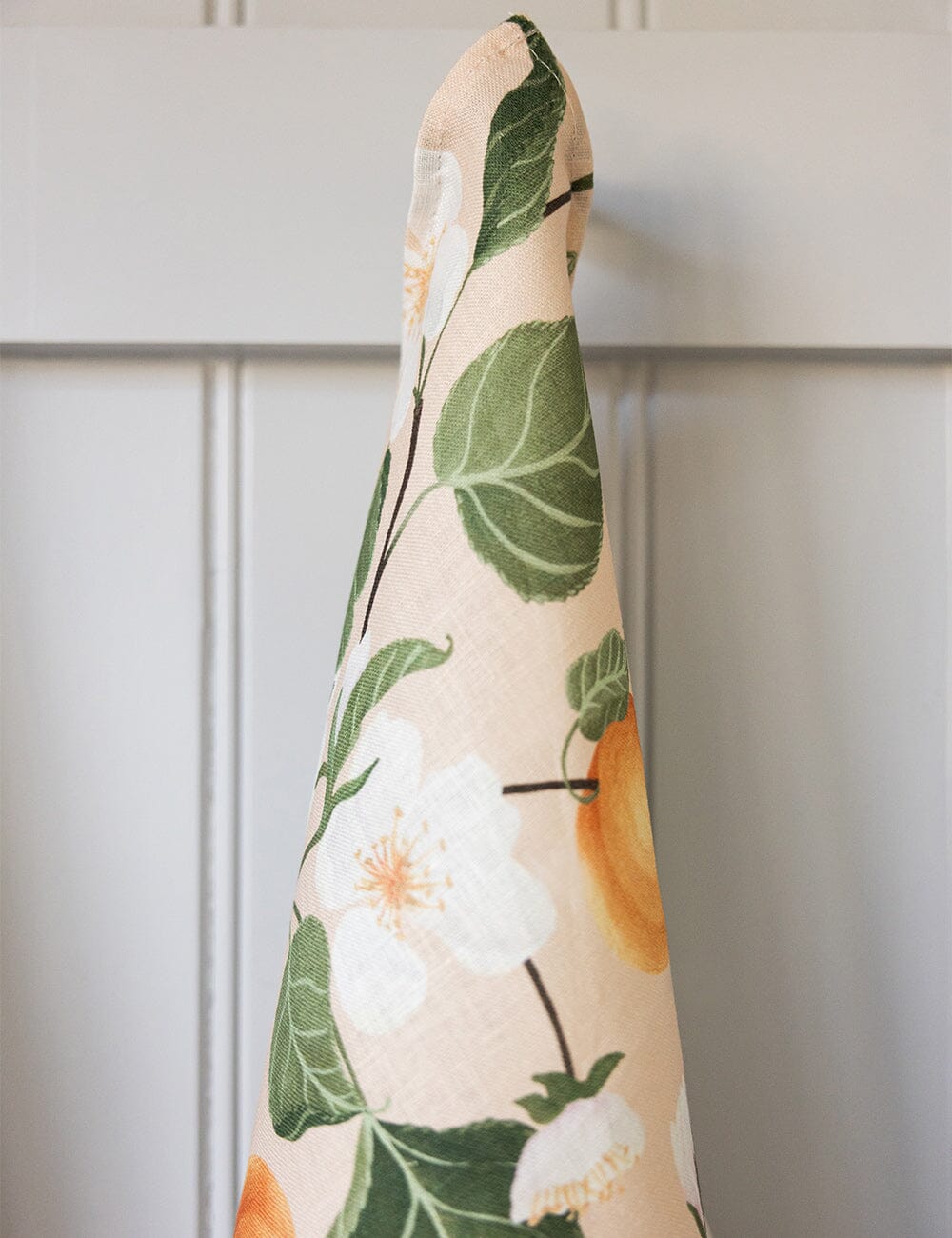 Close-up of the Peaches linen tea towel hanging from a hook, displaying hand-painted apricots, white blossoms, and green foliage on a soft blush background.