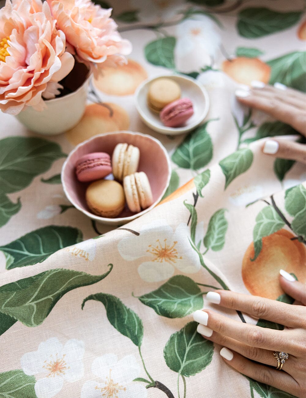 Peaches linen tea towel styled with blush peonies, pastel macarons, and delicate white floral illustrations, showcasing the fruit and leaf design on flax linen.