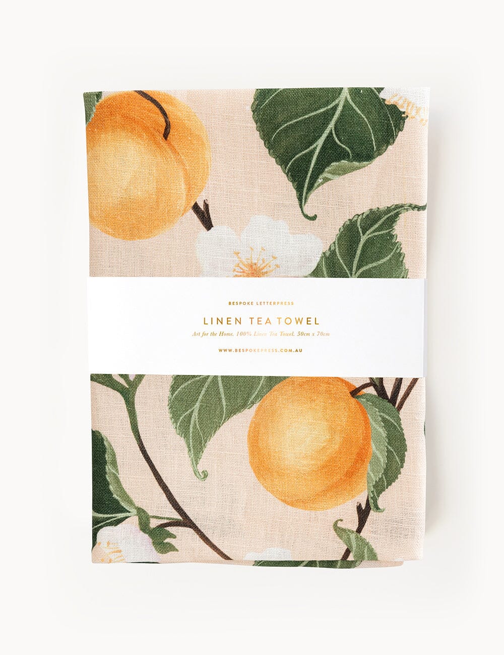 Peaches linen tea towel folded in packaging, featuring hand-painted apricot-toned fruit, white blossoms, and lush green leaves on 100% flax linen.