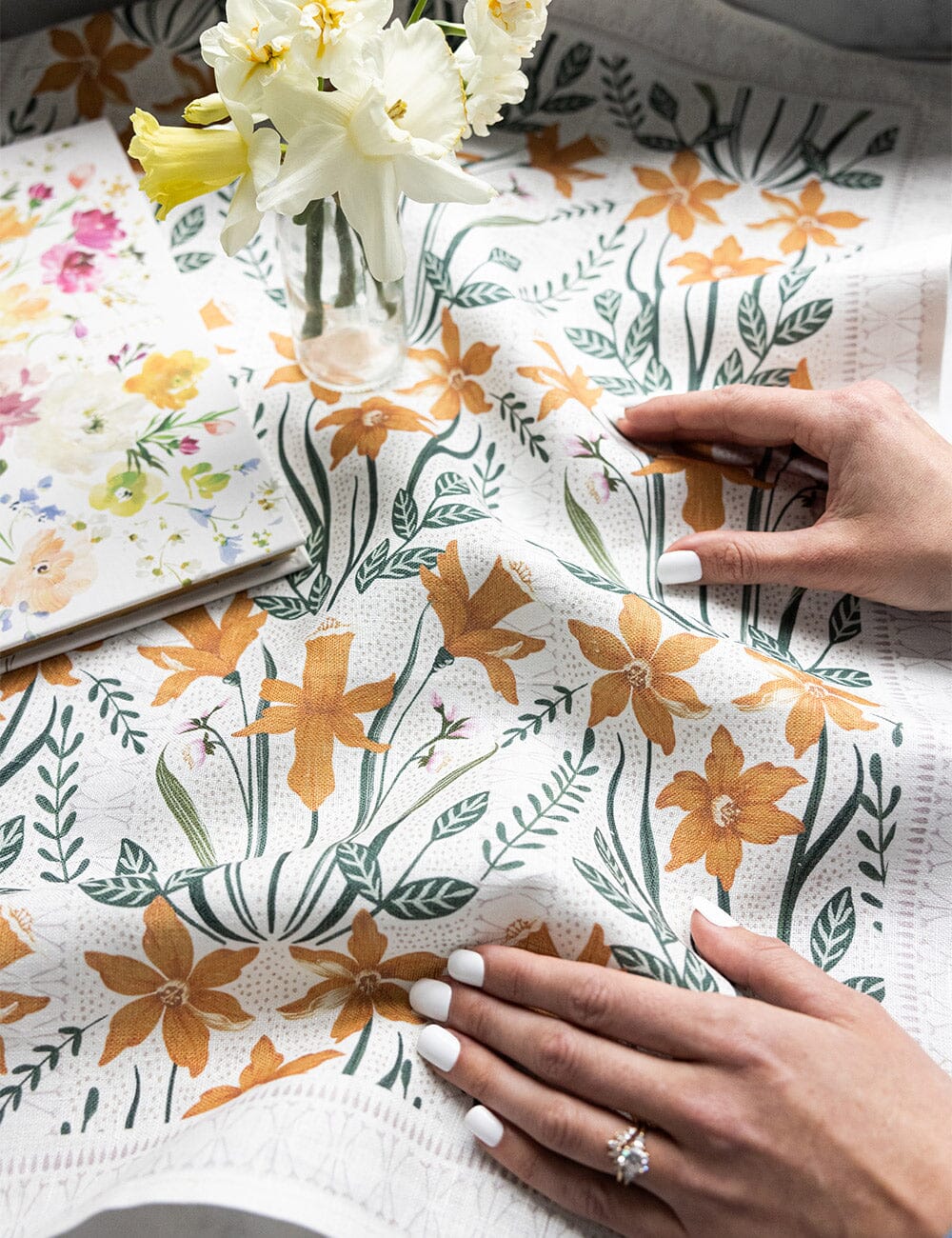 Close-up of hands gently smoothing a Bespoke Letterpress linen tea towel adorned with golden daffodils and green foliage, beside a vase of fresh daffodils and a floral notepad.