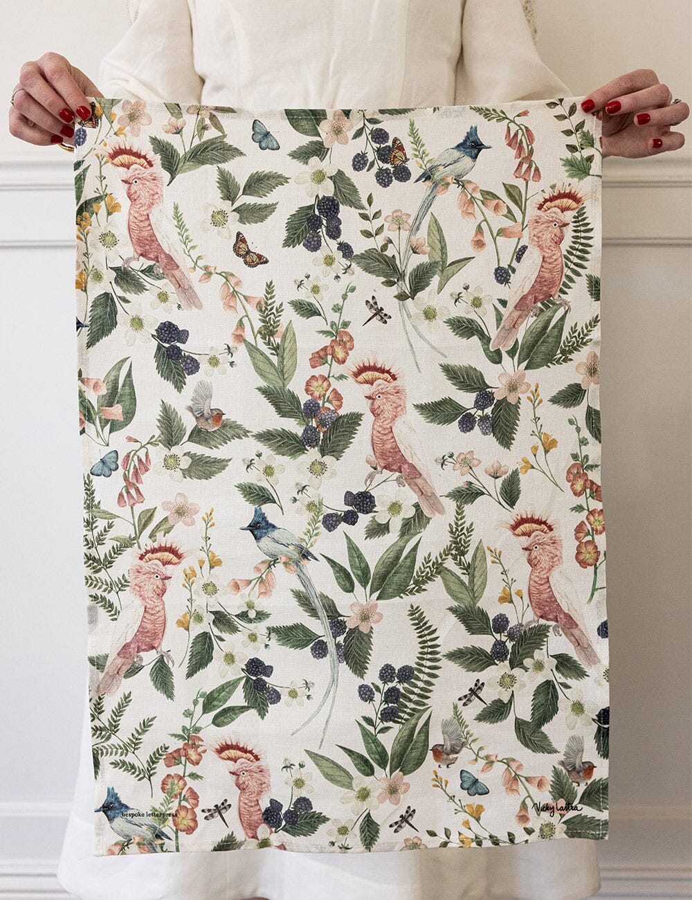 Person holding up the Pink Cockatoo linen tea towel, showcasing the full pattern of native birds, blackberries, butterflies, and Australian botanicals printed on natural flax linen