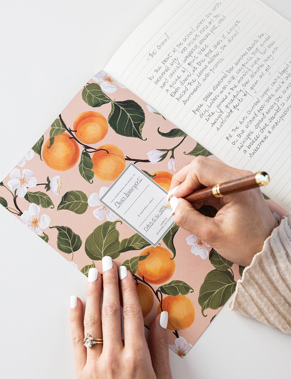 A hand writing on the nameplate inside the Peaches Workbook by Bespoke Letterpress, with lined pages open to handwritten notes and a backdrop of juicy peach illustrations and white blossoms on a blush-toned cover.