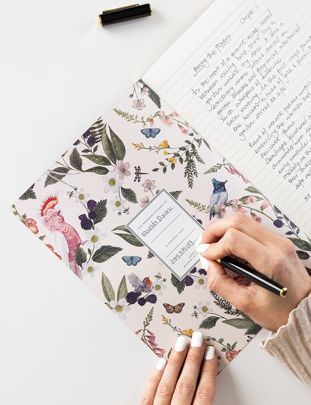 Person writing their name and contact details inside the Pink Cockatoo Workbook, which features a delicate Australian botanical and bird illustration on the inner cover, with lined pages open for journaling.