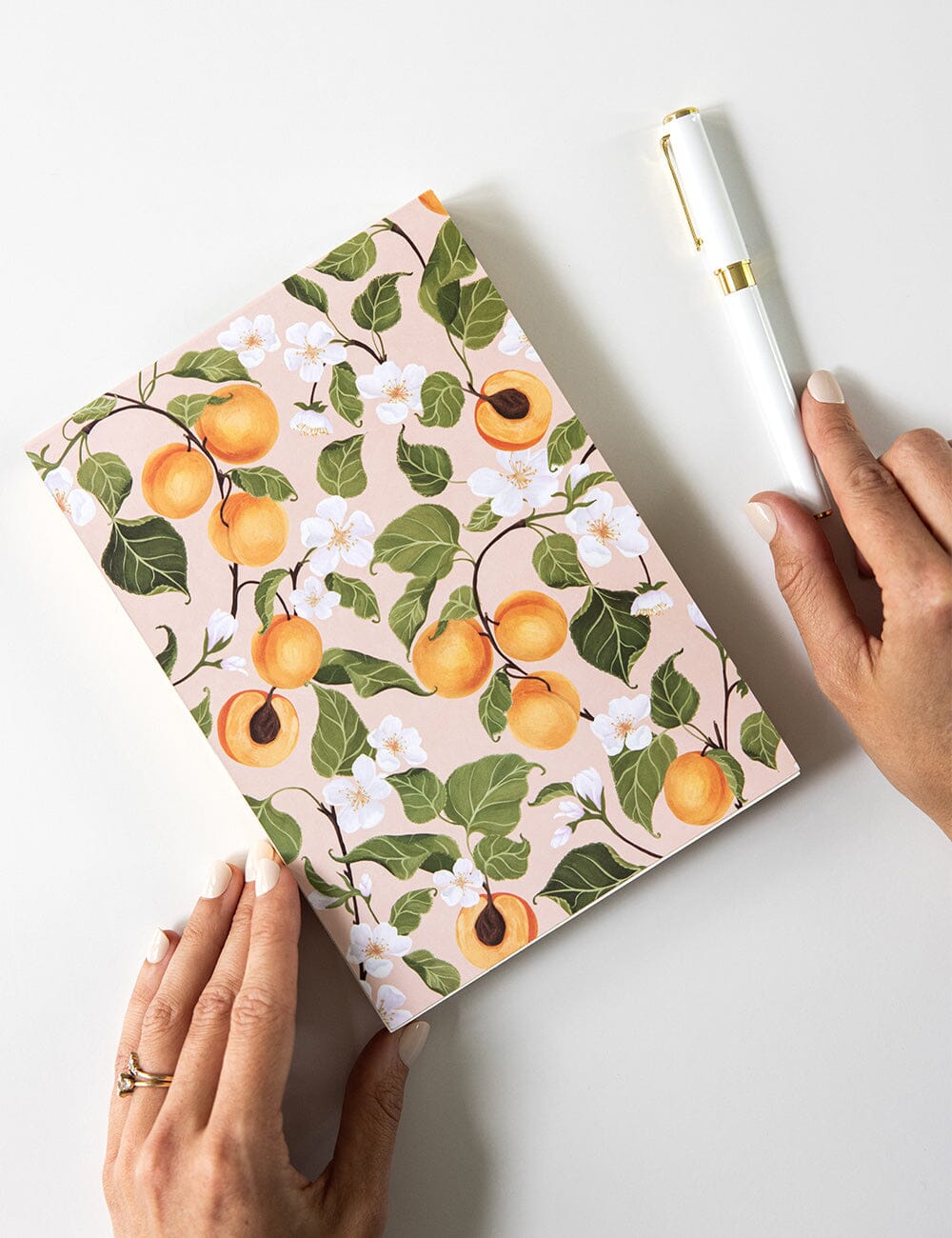 Woman holding a peach-themed A5 notepad jotter with botanical illustrations of ripe peaches and white blossoms on a blush background, paired with a white and gold ballpoint pen on a clean white desk.