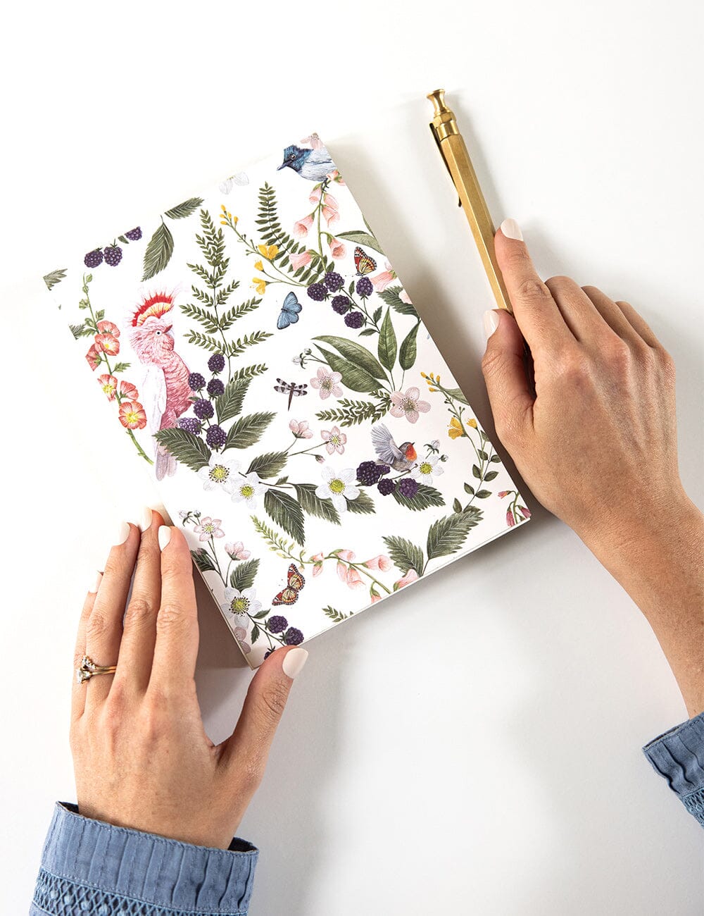 Hands holding the Pink Cockatoo Notepad Jotter by Bespoke Letterpress, featuring a botanical and bird illustration cover by Vicky Lastra, with a gold pen resting nearby on a white desk.