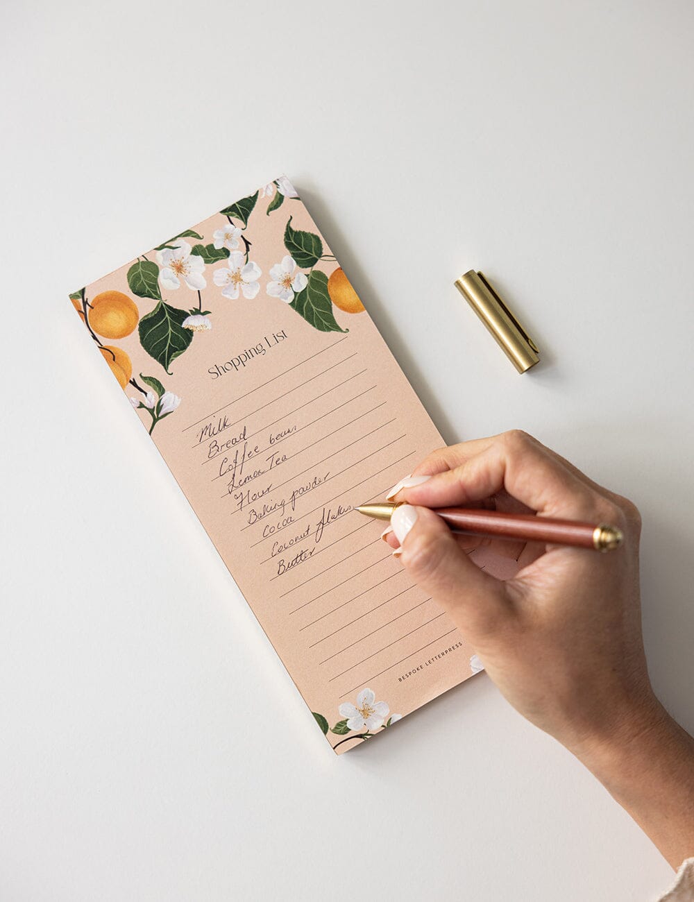 A hand writing on the Peaches Shopping List DL Notepad by Bespoke Letterpress, adorned with peach and floral illustrations on a soft blush background, featuring neatly lined pages for jotting down groceries with a gold pen placed nearby.
