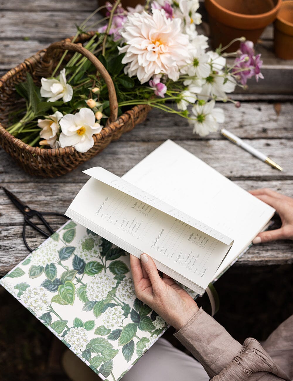 A person gently flips through the pages of the Bespoke Letterpress Gardening Journal, nestled on their lap beside a rustic timber table adorned with a wicker basket brimming with freshly cut blooms including dahlias, cosmos, and garden roses