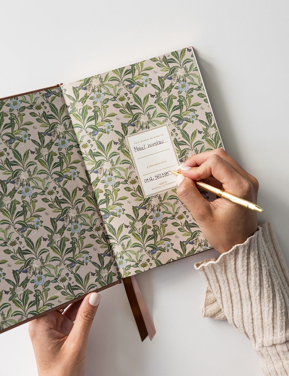 A hand holding open the front cover of the Journal to reveal a beautifully botanical green patterned print featuring a label with space to write your name. A hand is posed to write holding a gold pen.