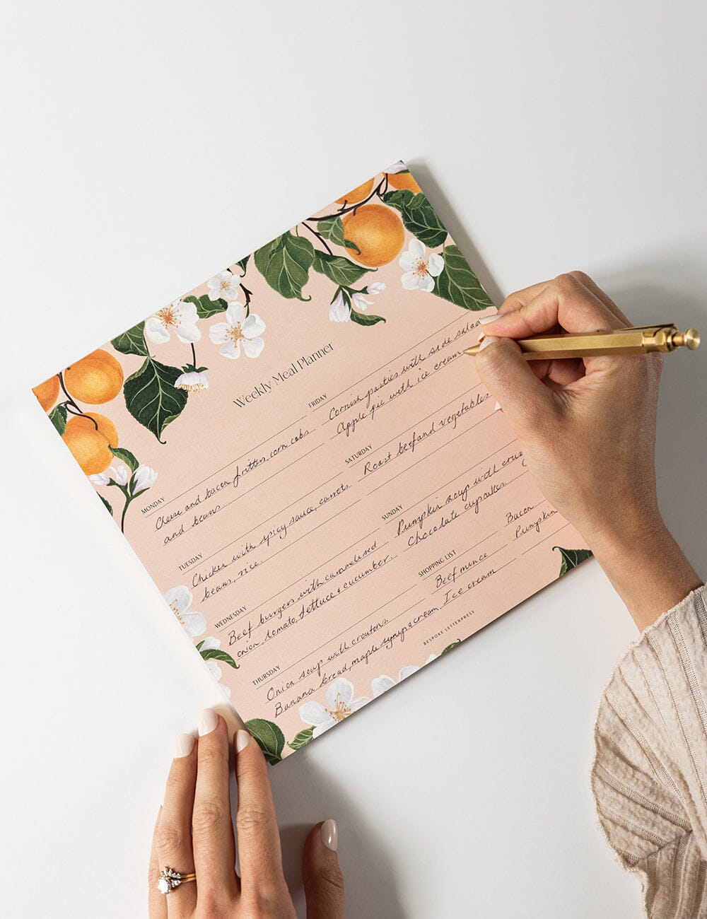 Hands writing on Bespoke Letterpress’ Peaches Meal Planner Notepad, featuring hand-illustrated peaches and blossoms with a structured weekly meal layout and shopping list section.