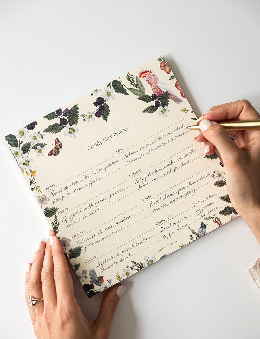 Hands writing on the Bespoke Letterpress 'Meal Planner Notepad – Cockatoo' filled with handwritten meal plans; features botanical and bird illustrations with a cream background and gold pen.