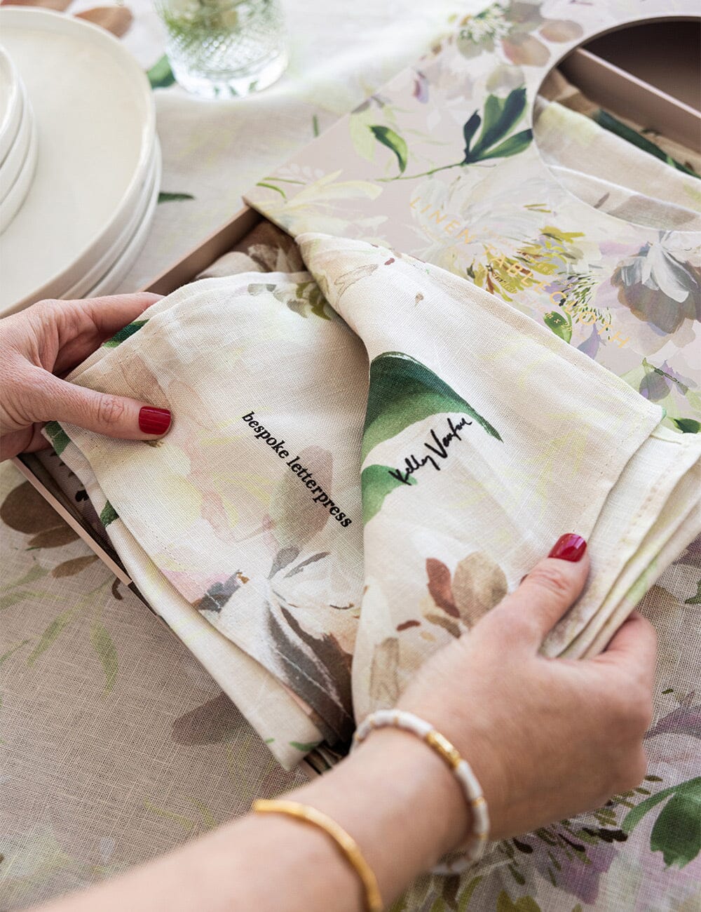 Hands unveiling the Peony Study linen tablecloth from its gift box, highlighting the floral fabric and artist collaboration labels.