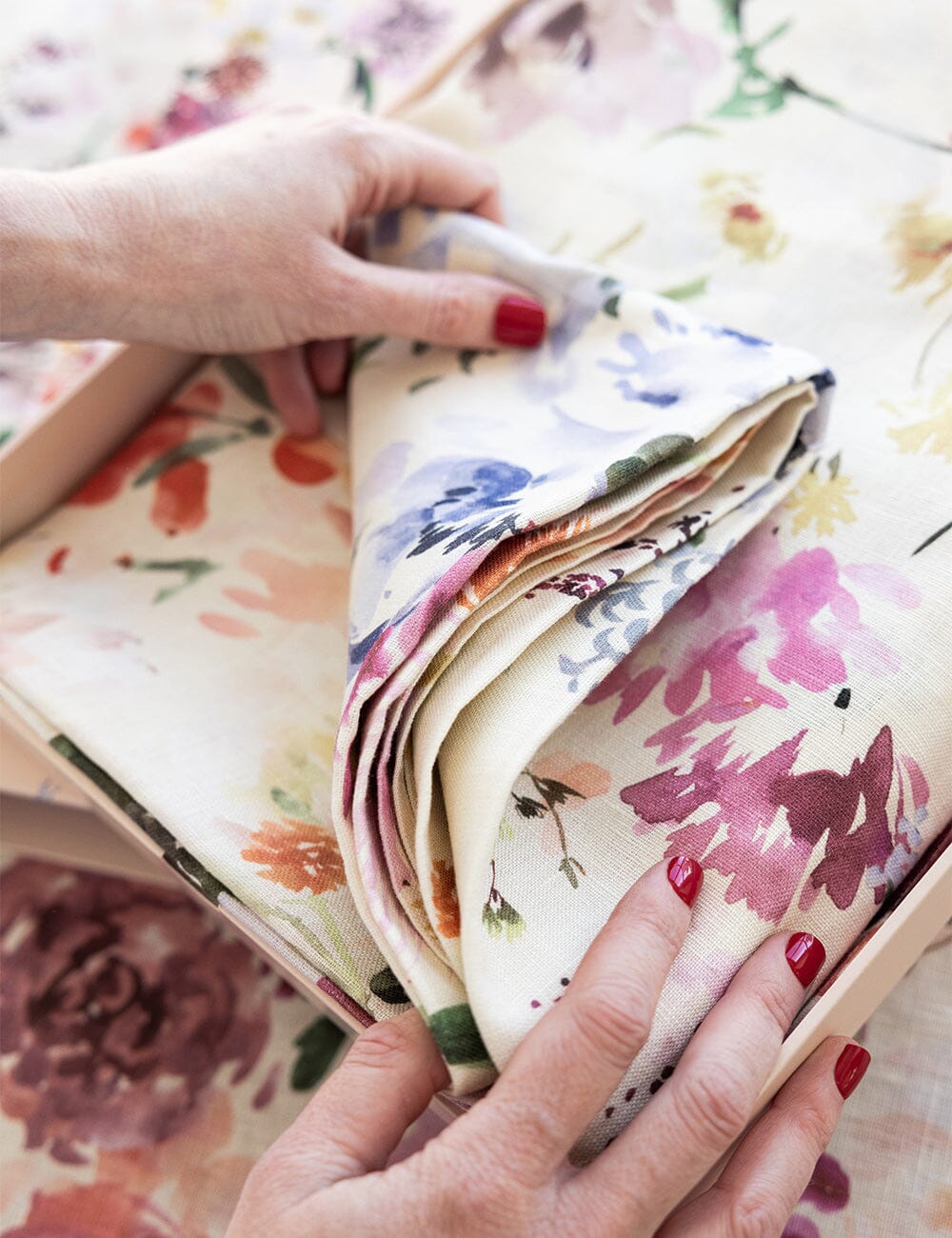 Hands unfolding the Wildflowers Linen Tablecloth, revealing its soft texture and painterly floral pattern in blush, violet, and golden hues.